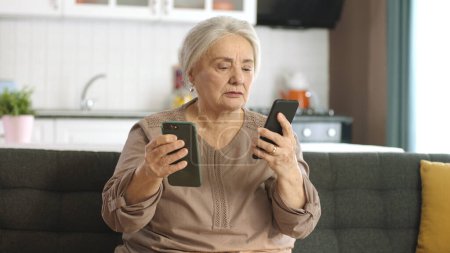 Téléchargez les photos : La vieille femme regarde ses comptes de médias sociaux, lit les nouvelles, écrit des messages sur ses deux smartphones. Une femme âgée qui essaie d'utiliser deux smartphones est surprise par ce qu'elle voit à l'écran. - en image libre de droit