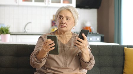 The old woman looks at her social media accounts, reads the news, writes messages on two smartphones. Elderly woman trying to use two smartphones talking to camera, looking at empty advertising space.