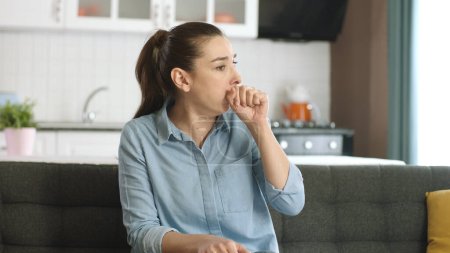 Eine junge Frau, die krank ist, hustet. Eine junge Frau in ihren Dreißigern hat im Wohnzimmer ihres Hauses Atembeschwerden. Atemnot, Asthma, Atemnot. Symptome des Coronavirus.