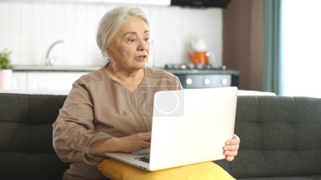 Senior mujer madura pasar el tiempo navegando por Internet en el ordenador portátil. Una mujer de 70 años está haciendo una videollamada en un chat virtual. Señora adulta de compras en línea en el sofá en la sala de estar.