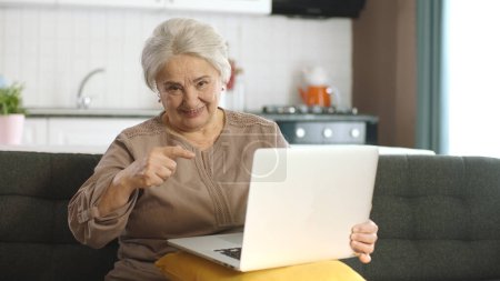 Elderly woman spends time surfing internet on laptop. Senior adult lady in her 70s shopping online on sofa in living room. Woman pointing finger at computer shows satisfaction with shopping.