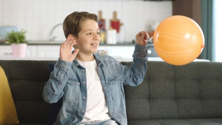 Lächelnder Junge, der allein im Wohnzimmer mit einem orangefarbenen Luftballon spielt, der für seine Geburtstagsfeier vorbereitet wurde. Ein einsamer, freundlicher, introvertierter, asozialer Junge blickt auf die rechte Seite des Bildschirms.