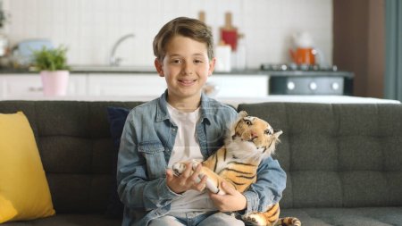 Allein zu Hause spielt ein freundlicher kleiner Junge mit Spielzeug-Tiger. Brauner Spielzeugtiger in Kinderhand. Junge spielt mit Spielzeug-Tiger im Wohnzimmer und lächelt in Kamera.