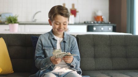 Schöner kleiner Junge sehr glücklich mit seinem Geld.Der Gewinn der Online-Lotterie. Der Junge macht aus Dollars einen Fan. Aufgeregter kleiner Junge genießt seinen Dollar allein zu Hause.