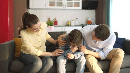 Ein junges Paar tröstet seinen traurigen kleinen Sohn im Wohnzimmer seines Hauses. Portrait von Eltern, die sich um ihre Kinder kümmern. Die Familie umarmt ihre Kinder, die in der Schule Probleme haben.