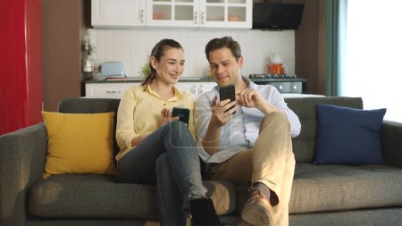 Téléchargez les photos : Couple heureux parlant riant sur le canapé en regardant smartphone à l'aide d'applications. Jeune couple magasinant en ligne, se réjouissant de l'argent qu'ils gagnent en ligne. - en image libre de droit