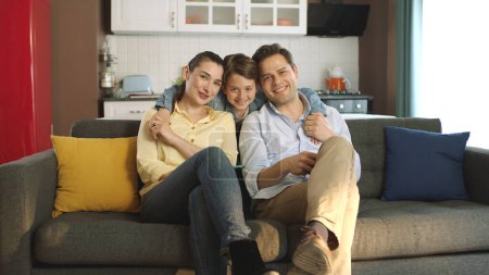 Téléchargez les photos : Portrait de famille heureuse assise sur un canapé dans leur maison paisible. Parents souriant à la caméra avec leur petit fils préscolaire mignon. - en image libre de droit