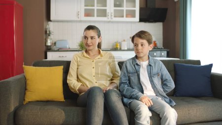 Junge Mutter und kleiner Junge sitzen mit Mutter auf Sofa im Wohnzimmer ihres Hauses. Er lächelt und blickt auf die leere Werbefläche links neben dem Bildschirm..