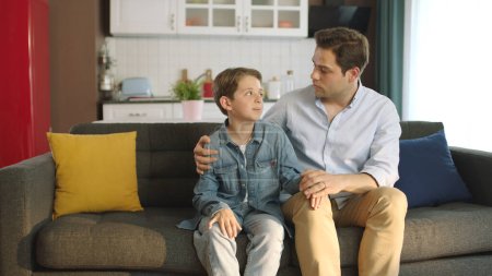 Téléchargez les photos : Portrait de jeune père et petit fils aimant leurs cheveux assis sur le canapé à la maison. Père réconfortant son petit fils qui a des problèmes à l'école ou dans la vie. Père réconfortant son fils triste. - en image libre de droit