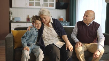 Foto de El niño visita a sus abuelos durante las vacaciones. Feliz pareja de ancianos sentados en el sofá y charlando con su nieto. Retrato de una familia feliz con nietos. - Imagen libre de derechos