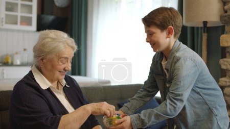 Téléchargez les photos : Garçon offrant des bonbons dans un bol à sa grand-mère âgée. Petit garçon offrant des bonbons à sa grand-mère âgée pour célébrer l'Aïd al-Fitr traditionnel (fête des bonbons) après la fin du Ramadan. - en image libre de droit