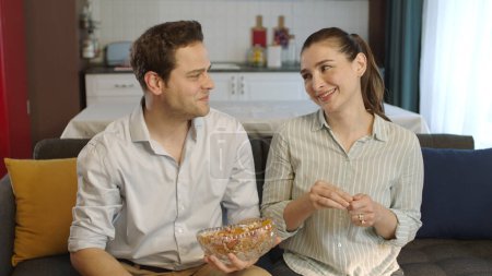 Photo for Young married couple loving each other eating mixed nuts. The woman and her husband, who eat hazelnuts, peanuts, almonds, watch movies on TV and have fun, laugh and are surprised at what they see. - Royalty Free Image