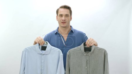 Photo for Young man isolated in front of white background holding two shirts. The man can't decide which shirt to wear. Not being able to decide on the choice of clothes. - Royalty Free Image