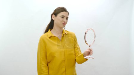 Téléchargez les photos : A woman holding a small mirror and checking her skin,hair.Young woman looking at her beauty,skin or hair problem in mirror isolated on white background surprised by what she sees.Beauty and skin care. - en image libre de droit