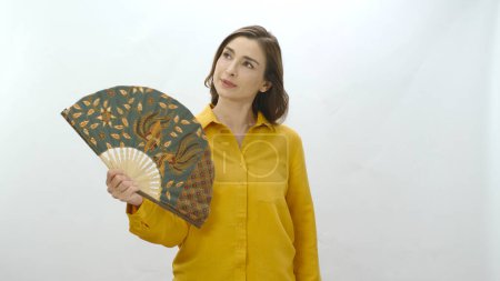 Photo for Young woman isolated on white background suffering from high temperature summer heat problem at home. Angry woman holding fan in hot weather looking at empty advertising space - Royalty Free Image