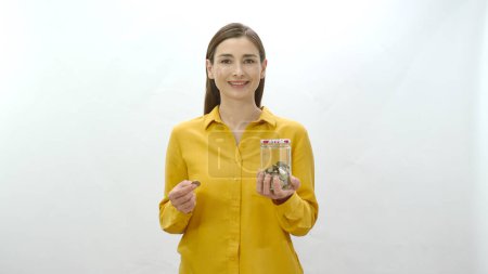 Charakterporträt einer jungen Frau, die gerne Geld spart, indem sie Geld in einem Glas anspart. Junge gesunde Frau blickt auf Sparschwein, sieht ihre Ersparnisse und fühlt sich glücklich.
