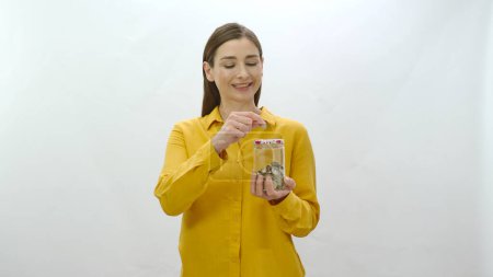 Charakterporträt einer jungen Frau, die gerne Geld spart, indem sie Geld in einem Glas anspart. Junge gesunde Frau wirft Münze in Sparschwein, sieht ihre Ersparnisse und fühlt sich glücklich.