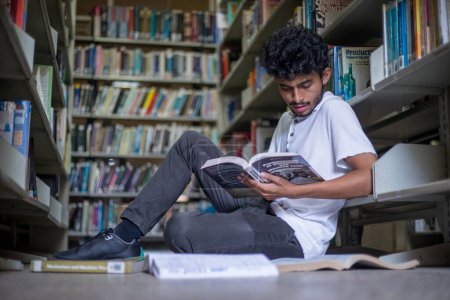 Photo for Young college student, book lover spending free leisure time in the library, mature man with a book in hands sitting on the floor near the bookcase, enjoying reading. Hobby concept - Royalty Free Image