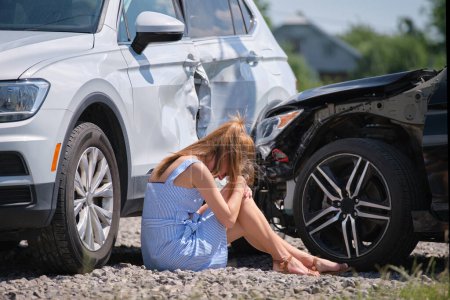 Photo for Sad young woman driver sitting near her smashed car looking shocked on crashed vehicles in road accident. - Royalty Free Image