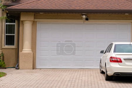 Foto de Vehículo estacionado en frente de una amplia puerta de garaje doble en la entrada pavimentada de la típica casa americana contemporánea. - Imagen libre de derechos