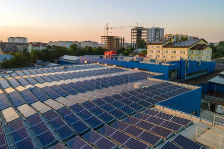 Aerial view of blue photovoltaic solar panels mounted on industrial building roof for producing green ecological electricity at sunset. Production of sustainable energy concept.
