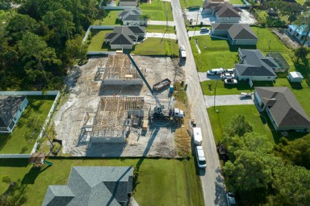 Lifting crane truck and builders working on roof construction of unfinished residential house with wooden frame structure in Florida suburban area. Housing development concept.