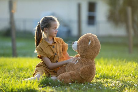 Photo for Pretty child girl tenderly embracing her teddy bear friend outdoors on green grass lawn. Concept of friendship values. - Royalty Free Image