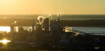 Luftaufnahme einer holzverarbeitenden Fabrik mit Rauch aus dem Produktionsprozess, der die Atmosphäre auf dem Werksgelände verschmutzt. Industriestandort bei Sonnenuntergang.
