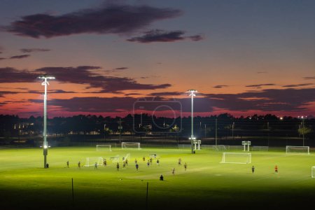Téléchargez les photos : Arène sportive publique éclairée à North Port, Floride avec des gens qui jouent au football sur le stade de football herbe au coucher du soleil. Concept d'activités extérieures. - en image libre de droit