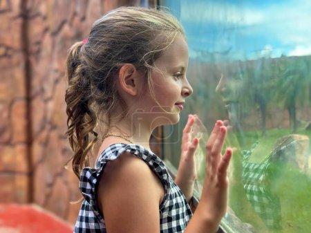 A young girl with braided hair in a checkered dress presses her hands against a window, looking out with a look of wonder.