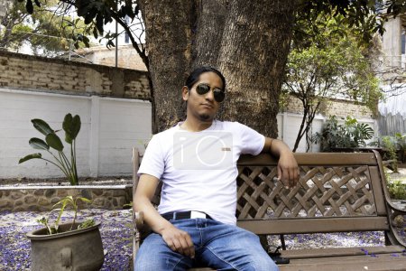 Foto de Hombre latino con camisa blanca y gafas de sol sentado en un banco - Imagen libre de derechos