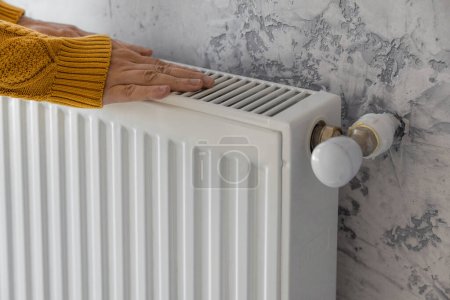 Foto de Man in yellow sweater warming his hands on the heater at home during cold winter days. Male getting warm up his arms over radiator. Concept of heating season or cold weather - Imagen libre de derechos