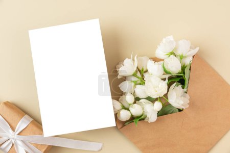 Blank greeting card with white jasmine flowers and gift box on beige background. Wedding invitation. Mock up. Flat lay. Mothers day layout