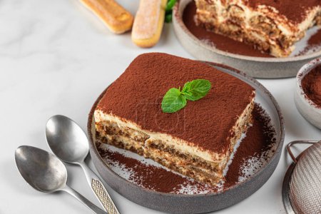 Téléchargez les photos : Dessert traditionnel italien tiramisu sur assiettes. Portions de gâteau au tiramisu maison avec menthe fraîche et cuillères sur table blanche. Nourriture savoureuse - en image libre de droit