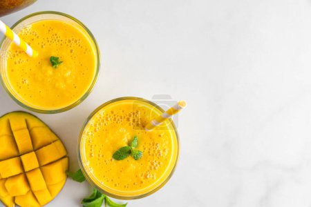 Mango smoothie in glasses with mint, fresh fruits and straw. Summer tropical drink on white background. Top view with copy space