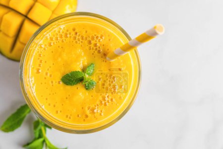 Téléchargez les photos : Smoothie à la mangue d'été jaune frais dans un verre avec une paille et menthe sur une table en marbre blanc. Vue de dessus. Boisson tropicale rafraîchissante - en image libre de droit