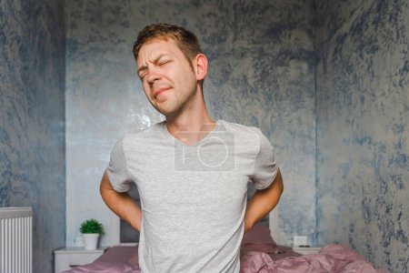 Photo for Young Caucasian man suffering from back pain while sitting on a bed at home. Kidney pain, backache, lumbago concept. Unhappy man with health problems - Royalty Free Image