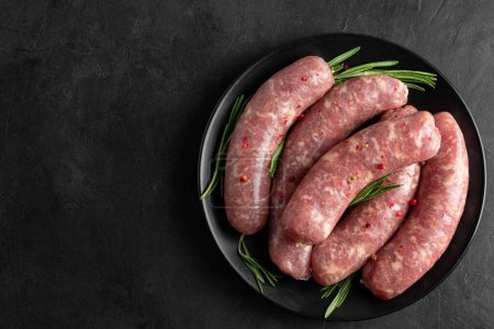 Raw sausages or bratwurst with spices and rosemary in a plate on black background. Top view. Food for grill or barbeque