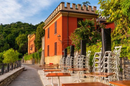 Cafétische draußen auf einem Damm mit farbenfroher Villa am Comer See in einem Außenrestaurant mit niemandem, Gemeinde Lenno, Lombardei, Italien. Beliebtes Reise- und Touristenziel im Sommerurlaub.