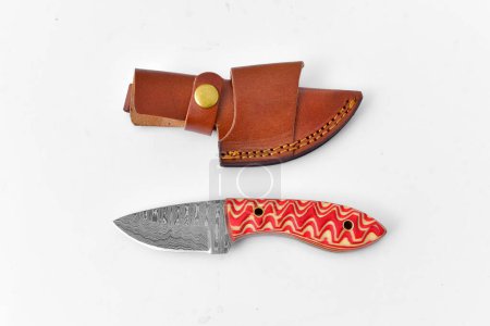Un pequeño cuchillo de hoja de Damasco con gran mango rojo anaranjado y funda de cuero sobre un fondo blanco.