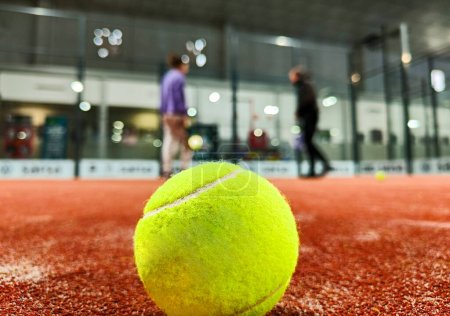 Téléchargez les photos : Plan détaillé d'une balle de paddle sur le sol du court de paddle-tennis intérieur et de trois amis joueurs en arrière-plan du court - en image libre de droit