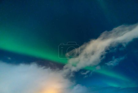 Photo for Aurora borealis with clouds illuminated by the full moon, glow of houses with a starry and bluish sky in Iceland. - Royalty Free Image