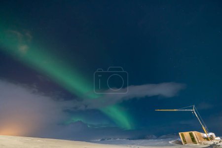 Photo for Aurora borealis with full moon lit clouds, glow of houses, construction crane and totally snowy ground with bluish starry sky in Iceland. - Royalty Free Image
