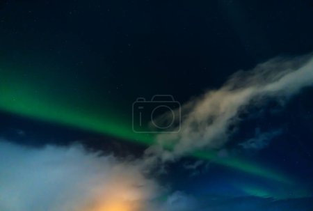 Photo for Aurora borealis with clouds illuminated by the full moon, glow of houses with a starry and bluish sky in Iceland. - Royalty Free Image