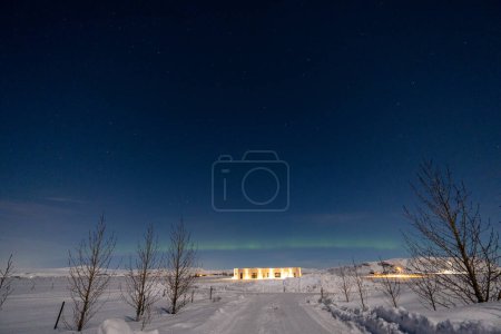 Photo for Northern lights on the horizon line and stars with a state of the art illuminated house at the end of a totally snowy road illuminated by moonlight in Iceland with tree silhouettes. - Royalty Free Image