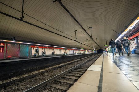 Photo for Train tracks and rails of the Barcelona metro or subway station and passengers waiting seated or walking with a train appearing from the beginning of the tunnel. - Royalty Free Image