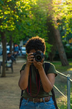 Téléchargez les photos : Joyeux jeune photojournaliste noir afro-américain aux cheveux bouclés foncés prenant des photos en regardant la caméra dans un parc vert au coucher du soleil avec l'anneau de mise au point coucher du soleil derrière - en image libre de droit