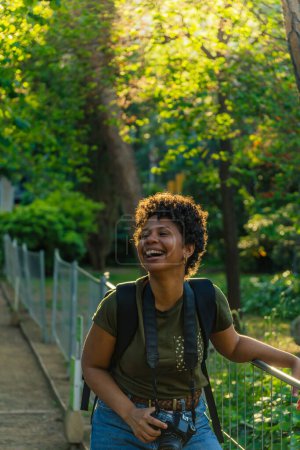 Téléchargez les photos : Joyeux jeune noir afro-américaine photojournaliste aux cheveux bouclés foncés rire heureux avec la bouche ouverte en regardant un ami avec appareil photo accroché autour du cou dans le parc vert au coucher du soleil - en image libre de droit