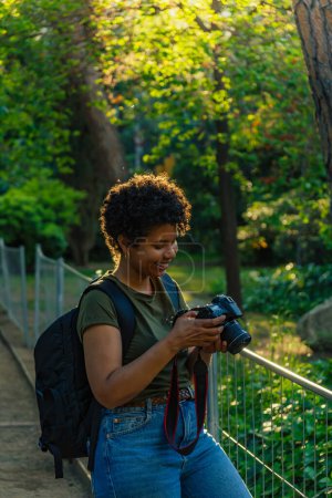 Téléchargez les photos : Heureux jeune noir afro-américain photojournaliste avec sombre cheveux bouclés vérification étonné et heureux avec bouche ouverte le résultat de photos prises avec un appareil photo numérique sur écran numérique dans le parc vert au coucher du soleil avec des paysages - en image libre de droit