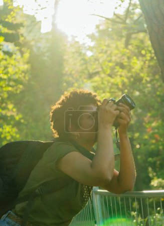 Téléchargez les photos : Jeune photojournaliste afro-américaine noire aux cheveux bouclés foncés prenant des photos dans un parc vert au coucher du soleil avec le soleil couchant derrière elle en ajustant la bague de mise au point de l'appareil photo - en image libre de droit